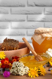Photo of Delicious honey, combs and different flowers on wooden table near white brick wall
