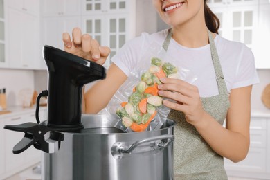 Photo of Woman putting vacuum packed vegetables into pot and using thermal immersion circulator in kitchen, closeup. Sous vide cooking