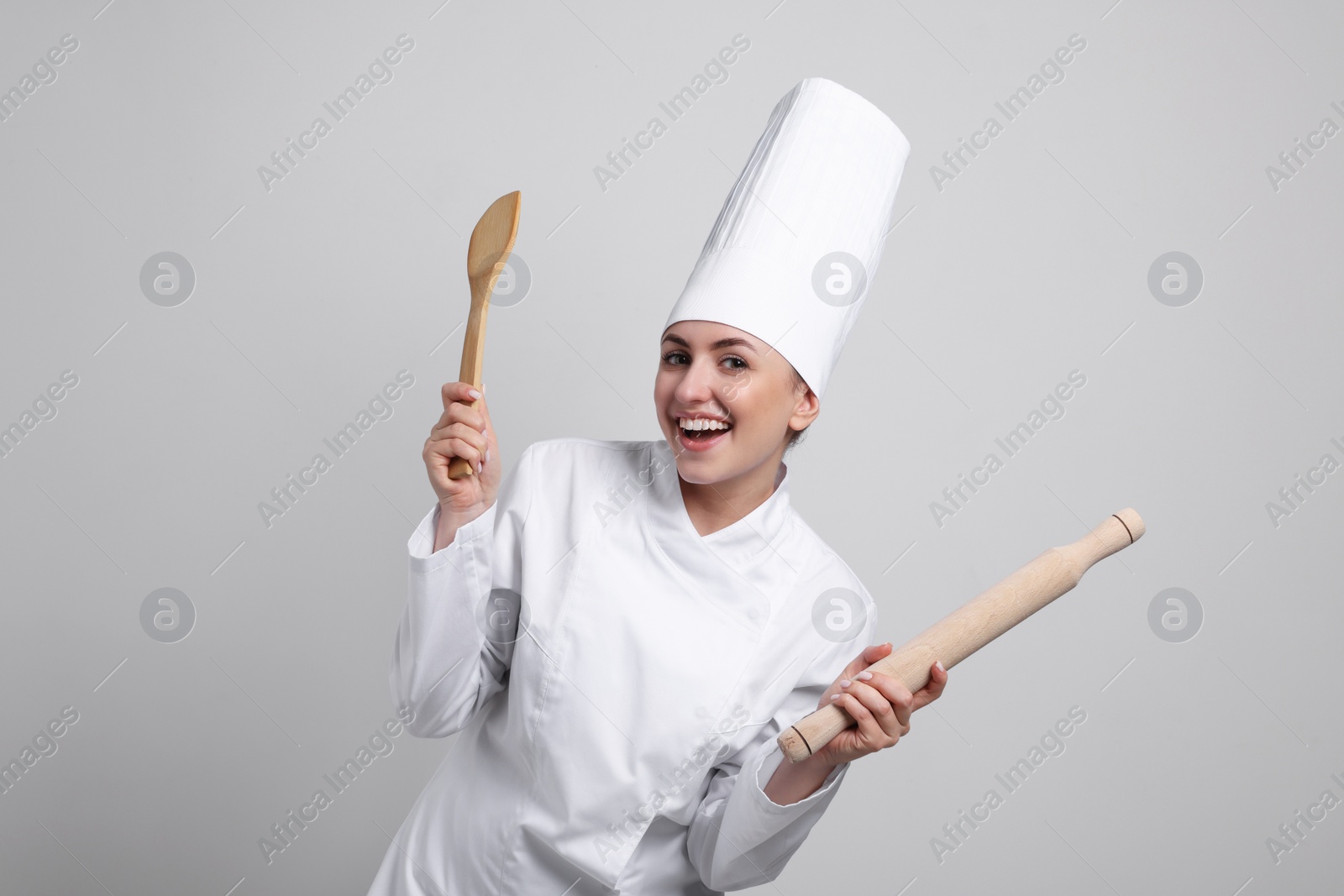 Photo of Happy professional confectioner in uniform holding wooden rolling pin and spatula on light grey background