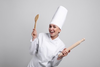 Happy professional confectioner in uniform holding wooden rolling pin and spatula on light grey background