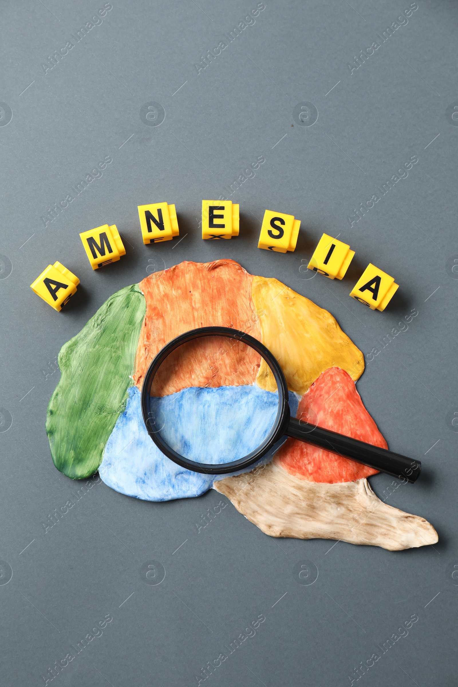 Photo of Word Amnesia made of yellow cubes, magnifying glass and brain with sections on grey background, flat lay