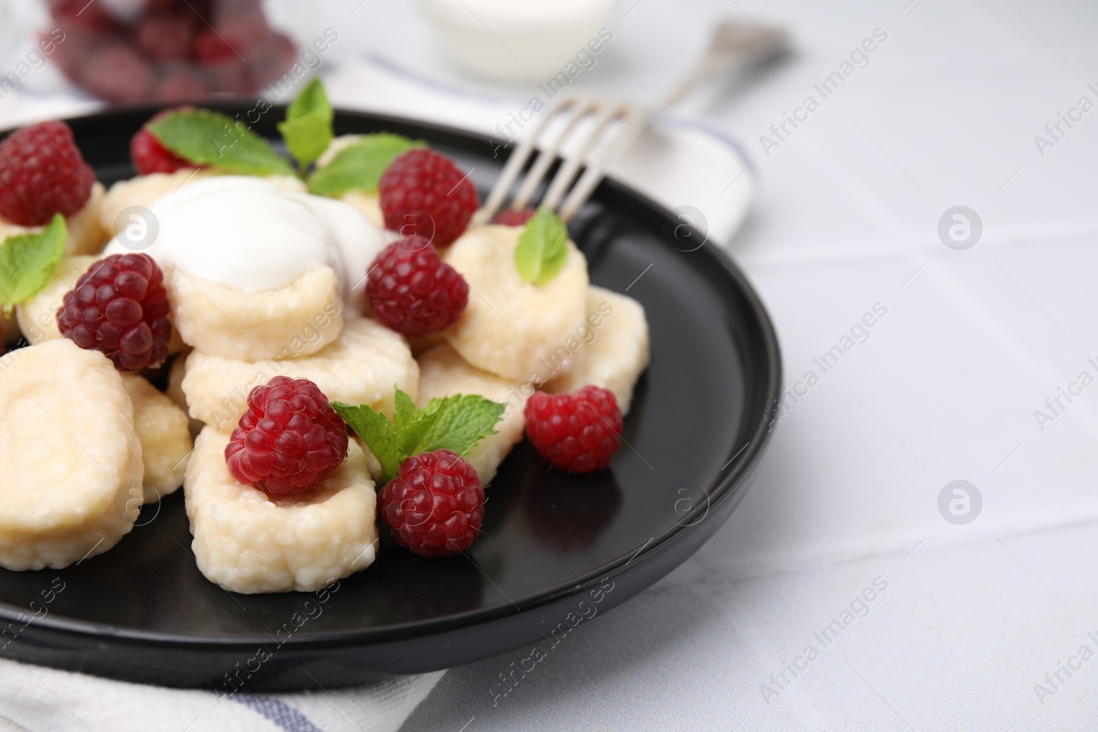Photo of Plate of tasty lazy dumplings with raspberries, sour cream and mint leaves on white tiled table, closeup. Space for text