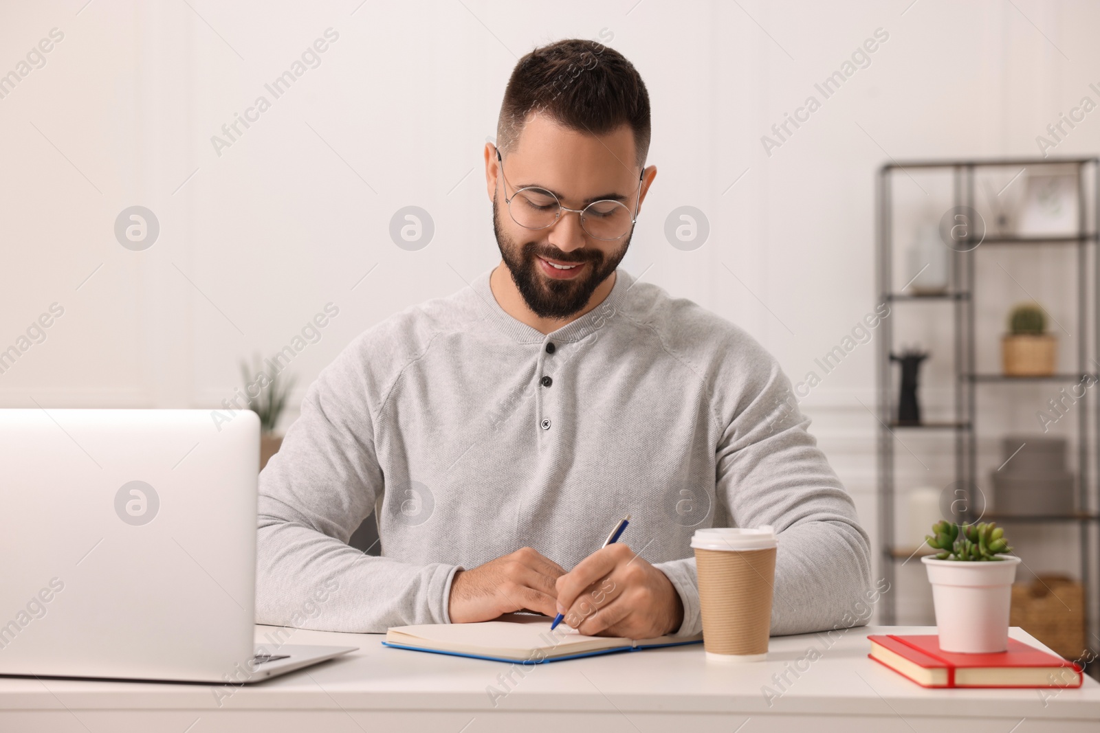 Photo of Young man writing in notebook at white table indoors