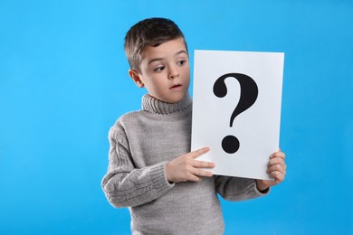 Emotional little boy holding paper with question mark on light blue background