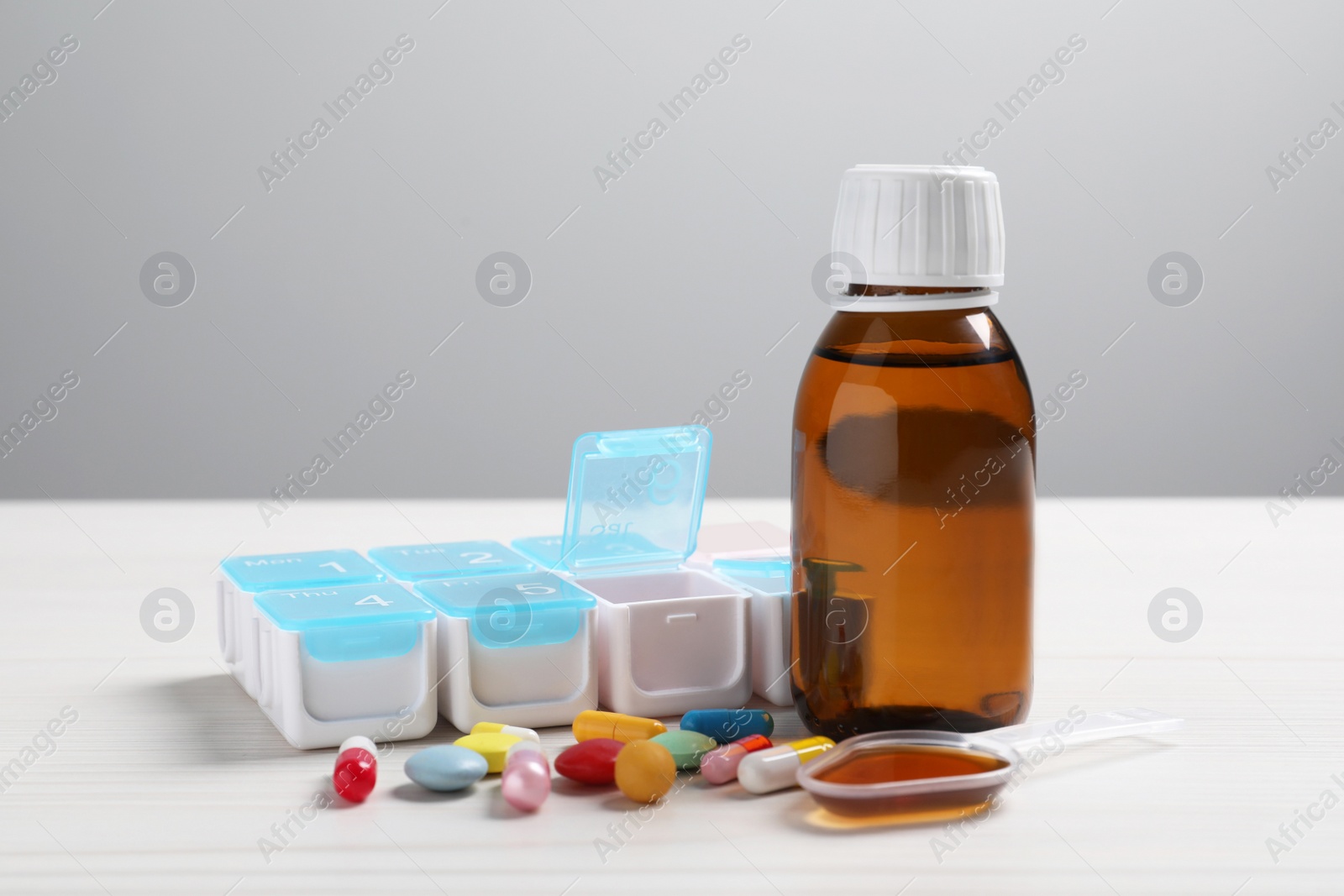 Photo of Bottle of syrup, dosing spoon and pills on white table against light grey background. Cold medicine
