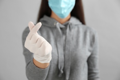 Woman in protective face mask and medical gloves showing korean heart gesture against grey background, focus on hand