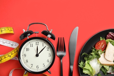 Photo of Plate of tasty salad, cutlery, alarm clock and measuring tape on red background, flat lay. Nutrition regime
