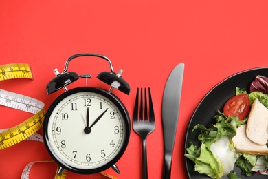 Plate of tasty salad, cutlery, alarm clock and measuring tape on red background, flat lay. Nutrition regime