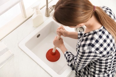 Photo of Young woman using plunger to unclog sink drain in kitchen, above view