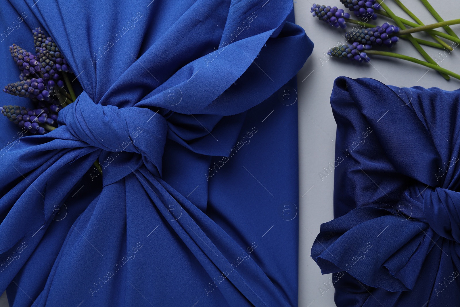 Photo of Furoshiki technique. Gifts packed in blue silk fabric and muscari flowers on white table, flat lay