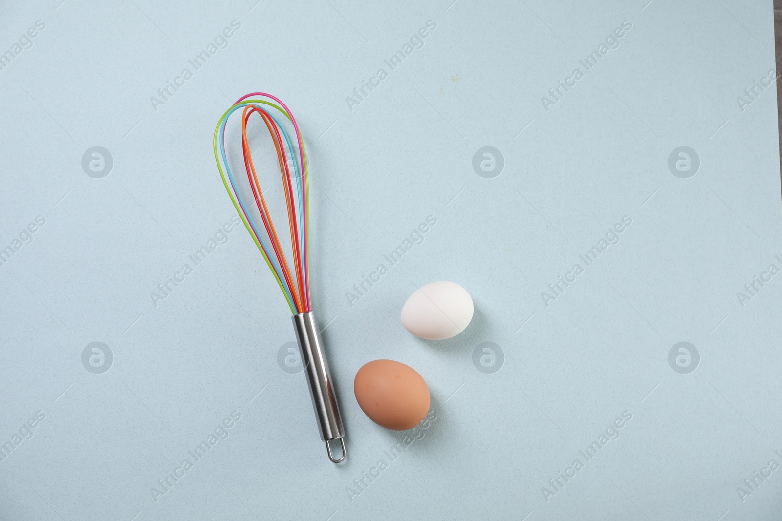 Photo of Colorful whisk and raw eggs on light blue background, flat lay