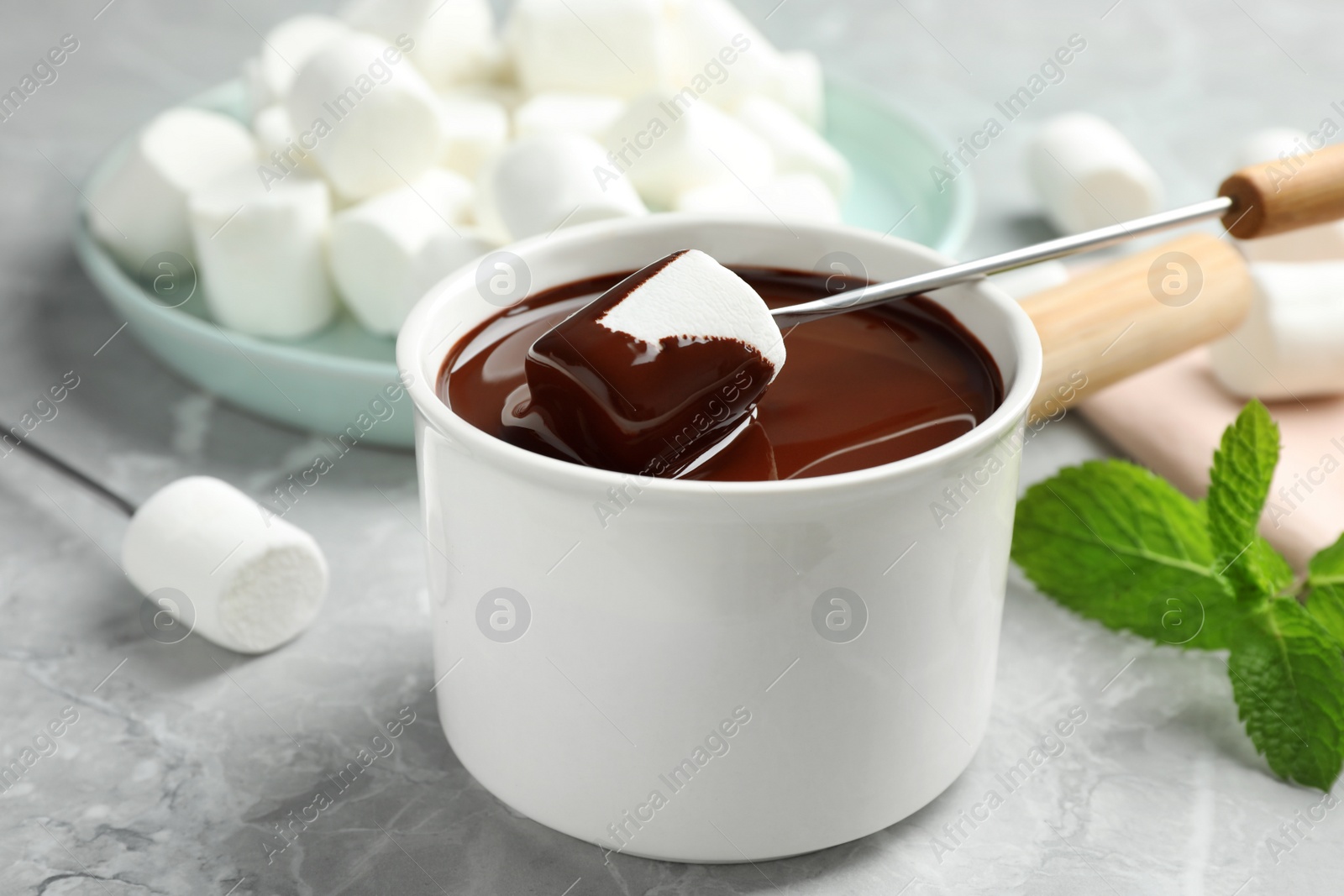 Photo of Dipping marshmallow into fondue pot with dark chocolate on marble table, closeup