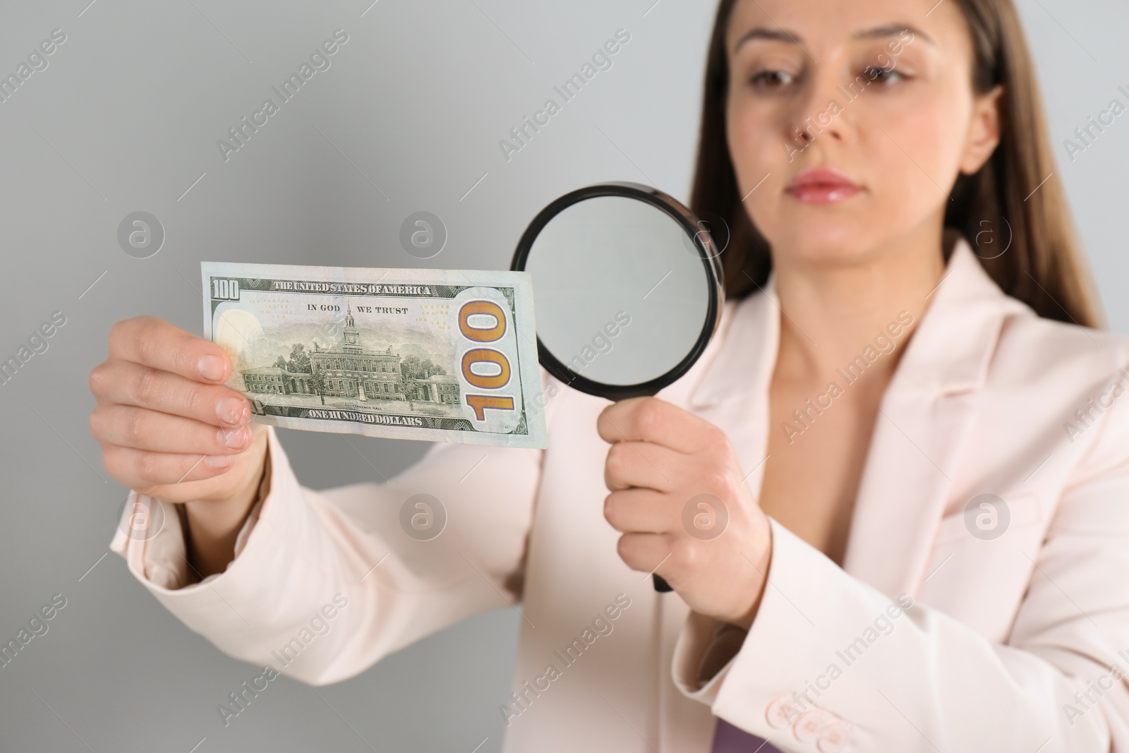 Photo of Expert authenticating 100 dollar banknote with magnifying glass against light grey background, focus on hand. Fake money concept