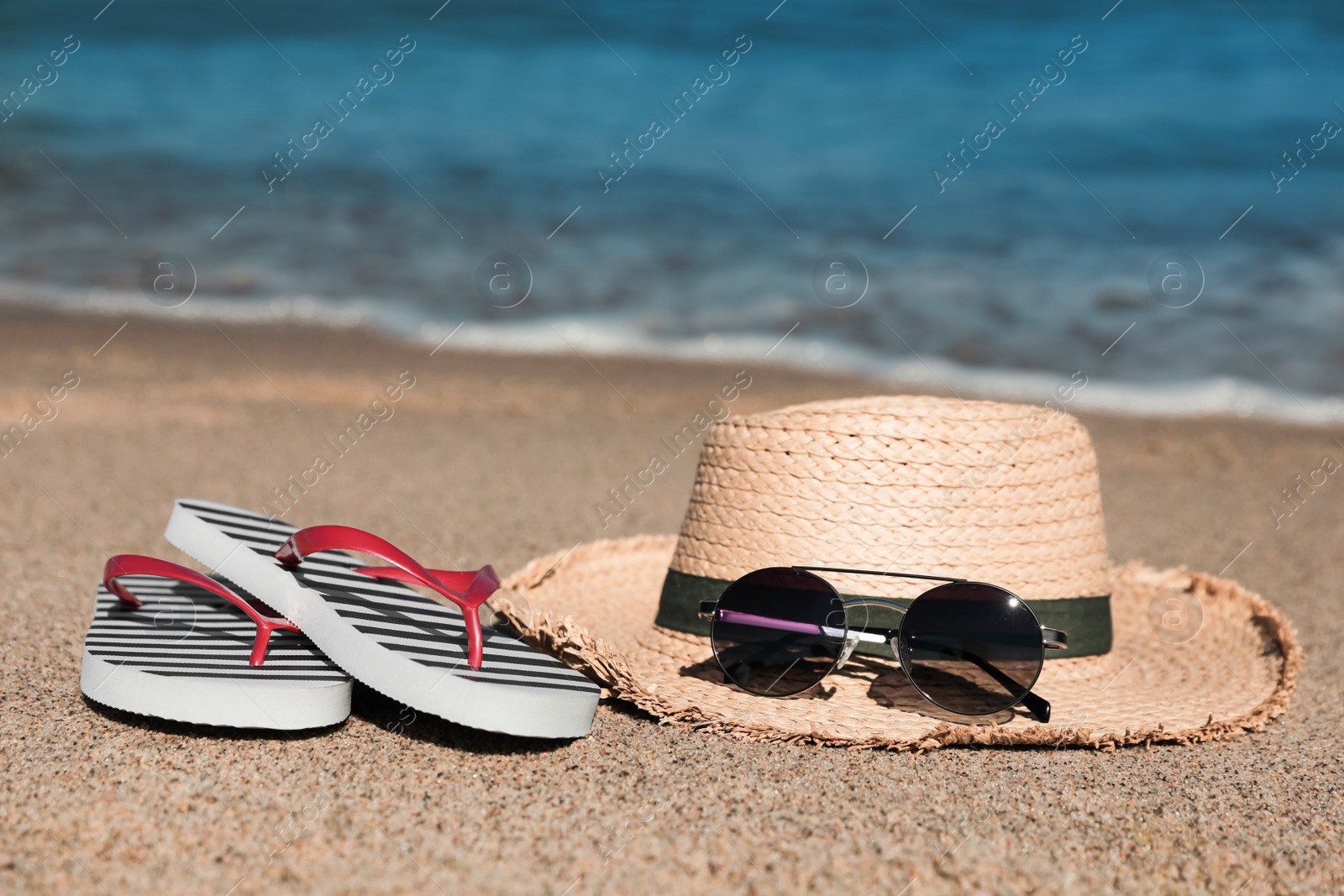 Photo of Striped flip flops, straw hat and sunglasses on sandy beach near sea