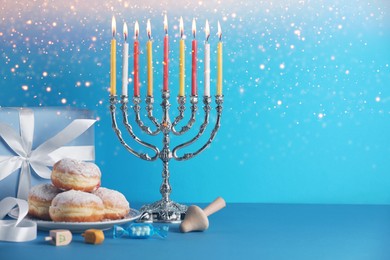 Image of Hanukkah celebration. Menorah with burning candles, dreidels, donuts and gift box on light blue table, space for text