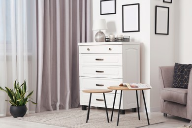 Empty frames hanging on white wall, chest of drawers and wooden tables indoors