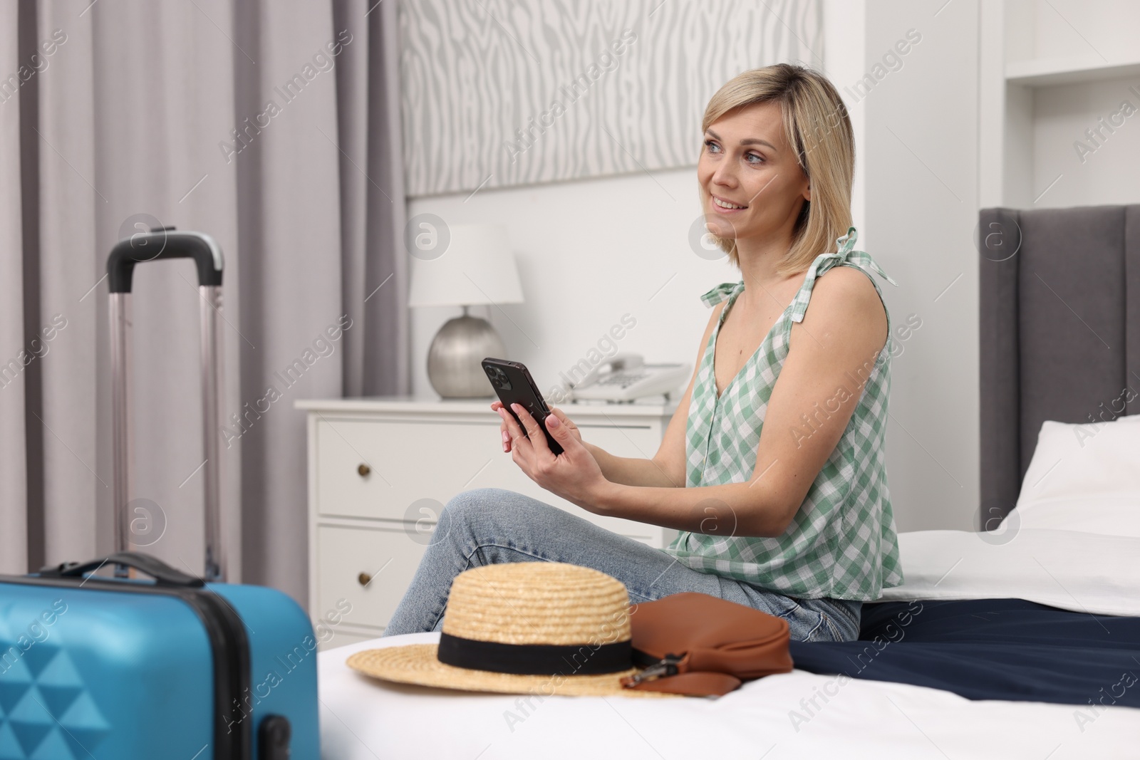 Photo of Smiling guest with smartphone relaxing on bed in stylish hotel room