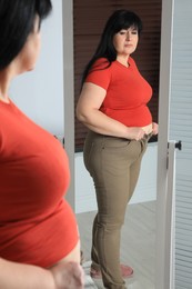 Overweight woman trying to button up tight trousers in front of mirror at home