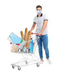 Young man in medical mask and shopping cart with purchases on white background. Coronavirus pandemic  