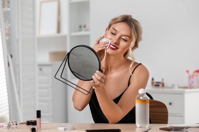 Photo of Smiling woman removing makeup with cotton pad in front of mirror at table indoors