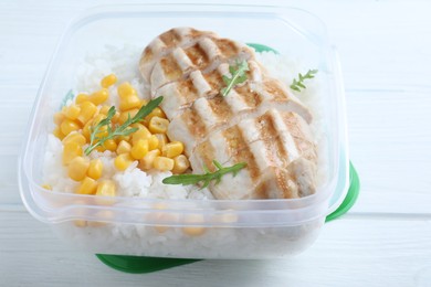 Tasty rice with grilled meat and corn in plastic container on white wooden table, closeup