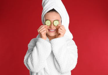 Young woman in bathrobe with cucumber slices on red background