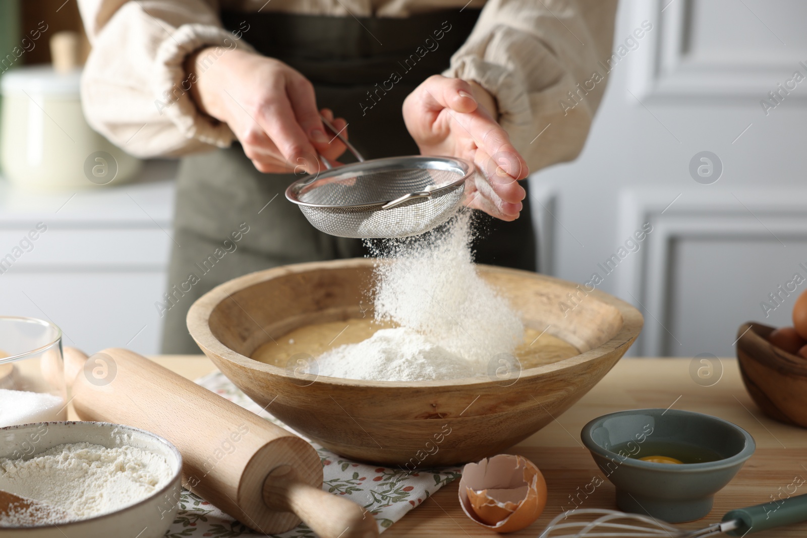 Photo of Making dough. Woman sifting flour into bowl at wooden table in kitchen, closeup