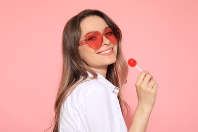 Photo of Portrait of beautiful young woman with heart shaped sunglasses and lollipop on color background