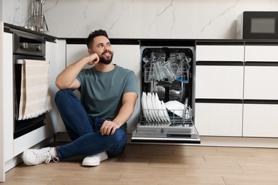 Photo of Smiling man sitting near open dishwasher in kitchen