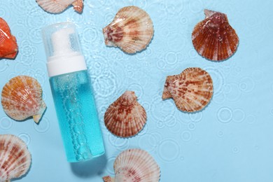 Bottle of face cleansing product and seashells in water against light blue background, flat lay. Space for text