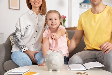 Photo of Happy family putting coin into piggy bank at table indoors. Saving money