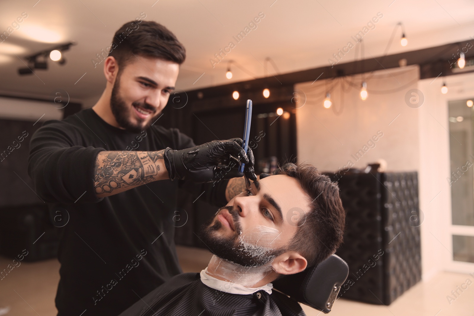 Photo of Professional hairdresser shaving client with straight razor in barbershop