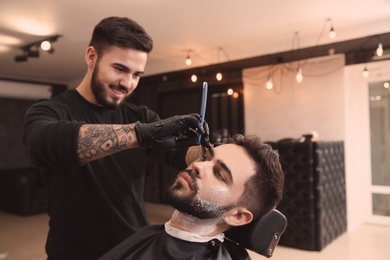 Photo of Professional hairdresser shaving client with straight razor in barbershop