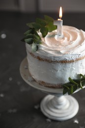 Tasty Birthday cake with burning candle and eucalyptus branches on grey table, closeup