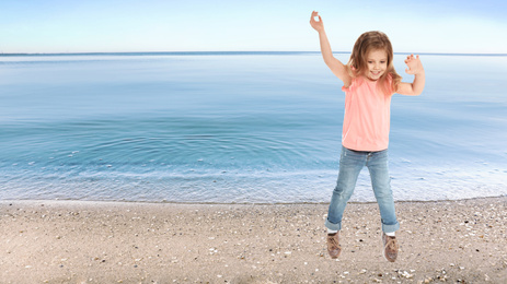 Happy school girl jumping on beach near sea, space for text. Summer holidays