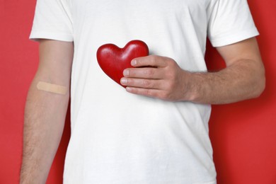 Blood donation concept. Man with adhesive plaster on arm holding red heart against color background, closeup