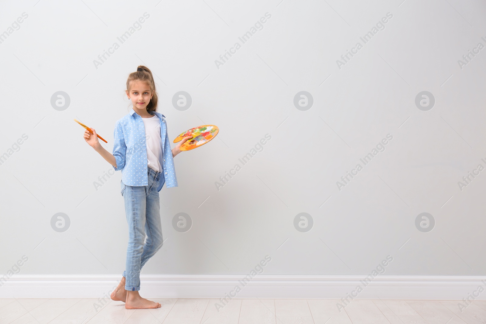 Photo of Little girl with brush and paint palette near light wall indoors. Space for text