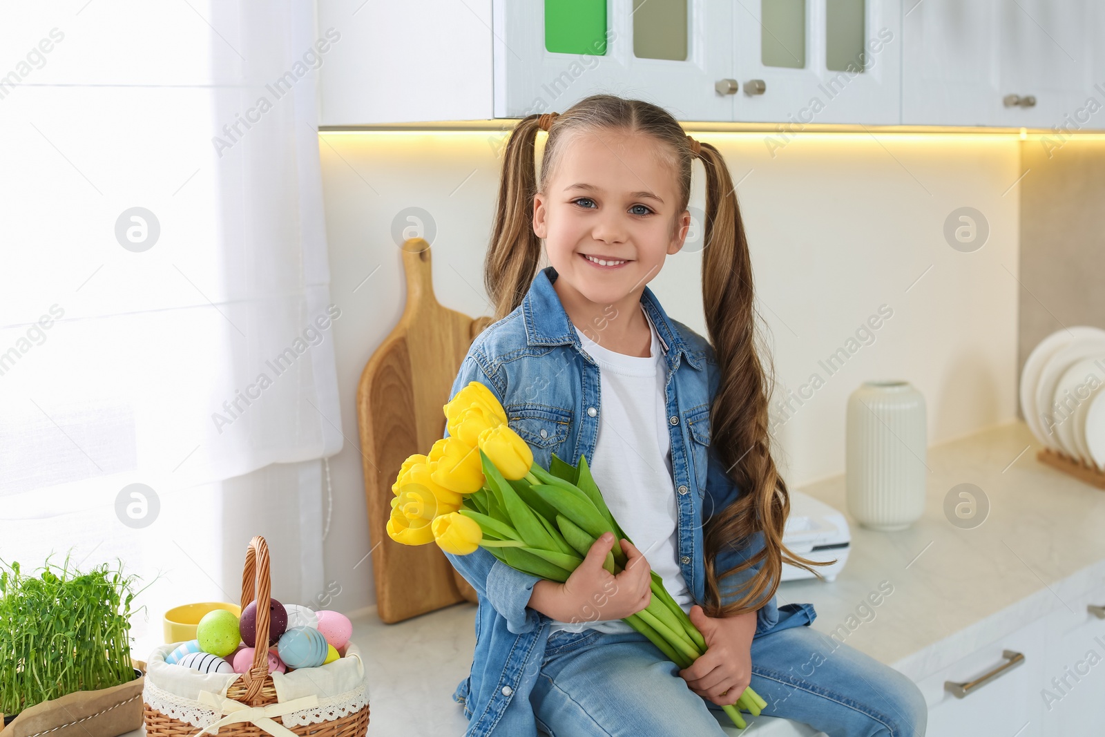 Photo of Cute girl with yellow tulips and wicker basket full of Easter eggs in kitchen