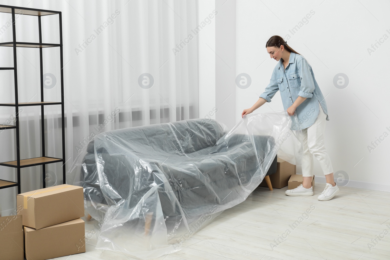 Photo of Young woman covering sofa with plastic film at home