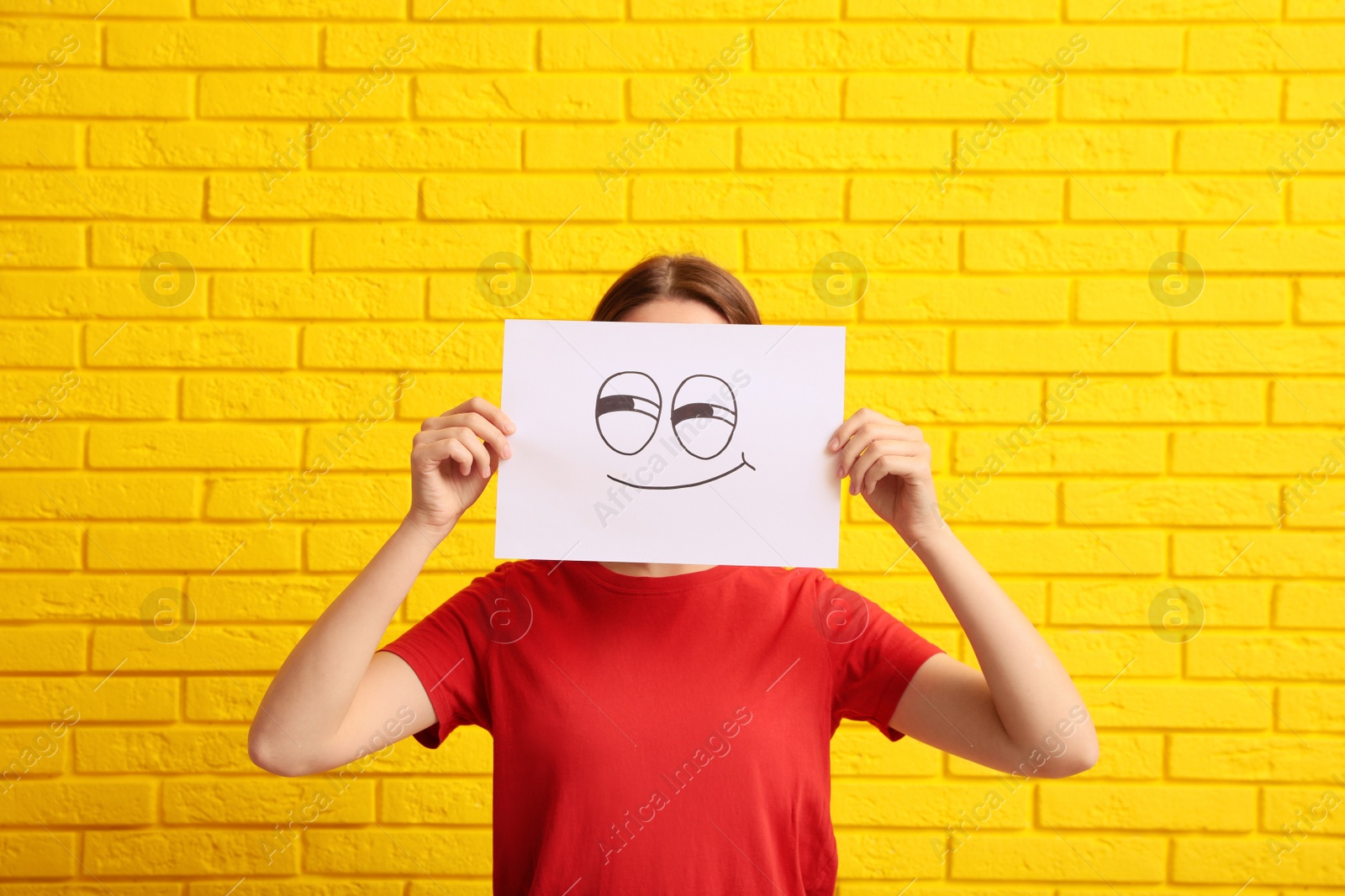 Photo of Woman hiding behind sheet of paper with smirking face near yellow brick wall
