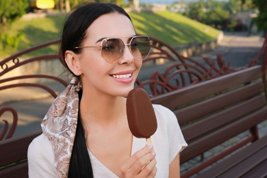 Photo of Beautiful young woman holding ice cream glazed in chocolate on bench outdoors