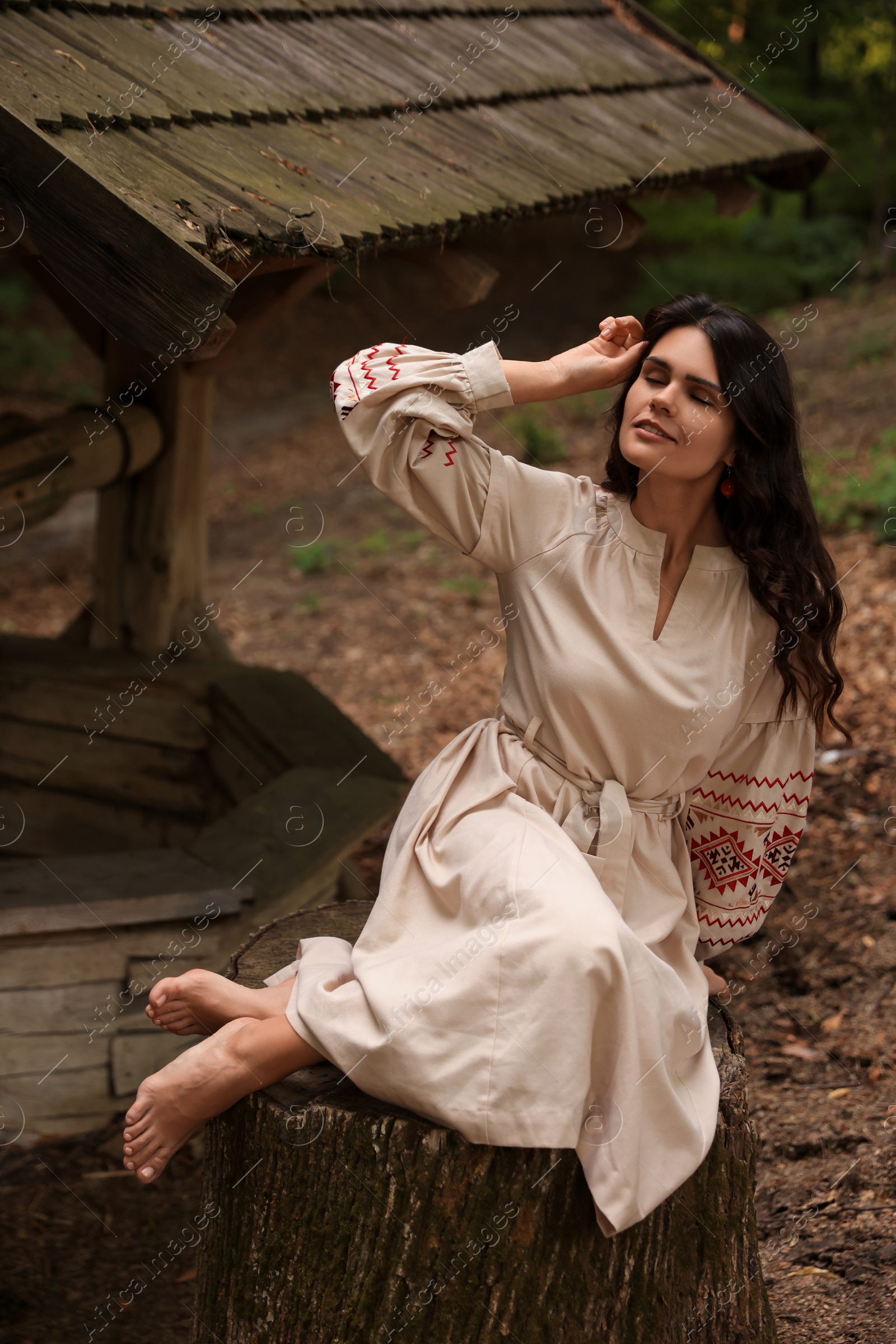 Photo of Beautiful woman wearing embroidered dress sitting near old wooden well in countryside. Ukrainian national clothes