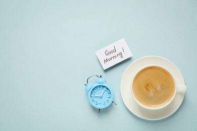 Photo of Delicious coffee, alarm clock and card with GOOD MORNING wish on light background, flat lay. Space for text