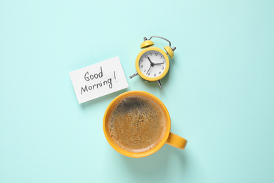 Photo of Message GOOD MORNING, alarm clock and coffee on light blue background, flat lay