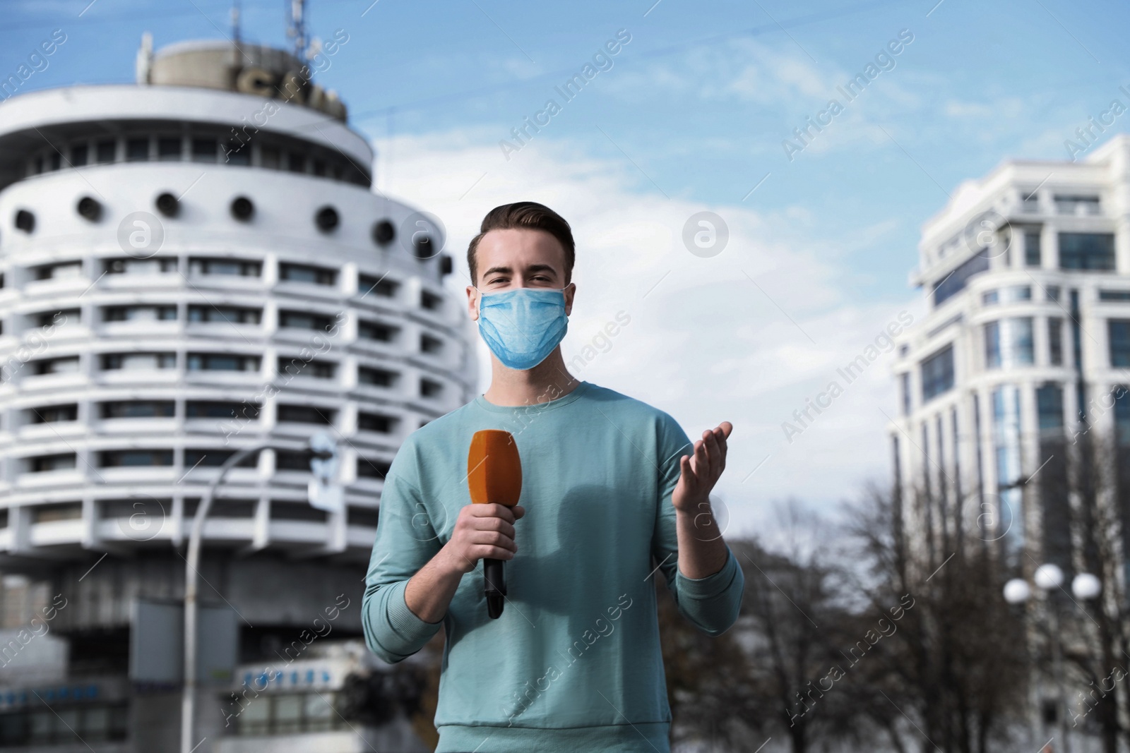 Image of Young journalist with medical mask and microphone working on city street. Virus protection