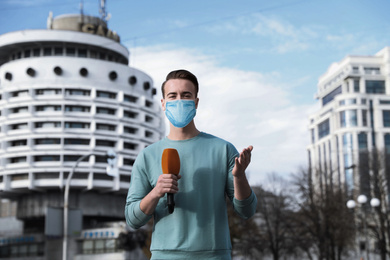 Young journalist with medical mask and microphone working on city street. Virus protection