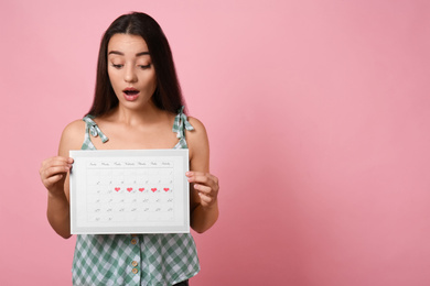 Photo of Young woman holding calendar with marked menstrual cycle days on pink background. Space for text