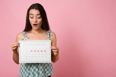 Young woman holding calendar with marked menstrual cycle days on pink background. Space for text