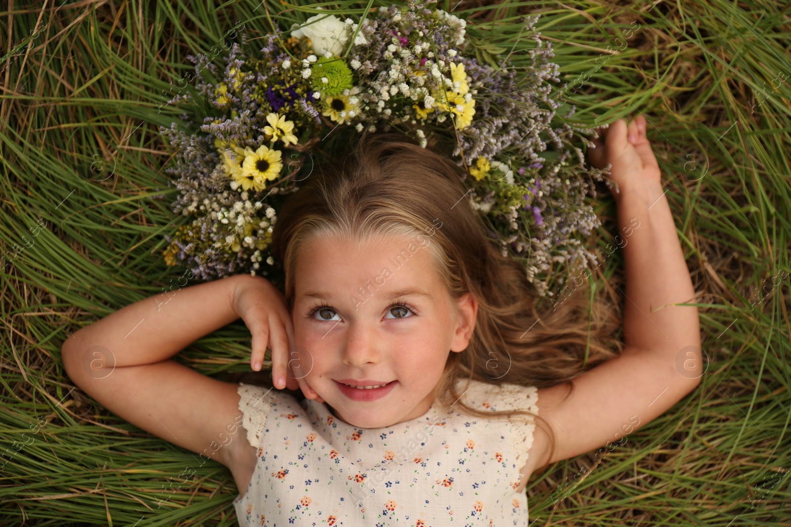 Photo of Cute little girl wearing wreath made of beautiful flowers on green grass, top view