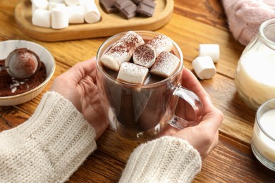 Photo of Woman holding cup of aromatic hot chocolate with marshmallows and cocoa powder at wooden table, closeup